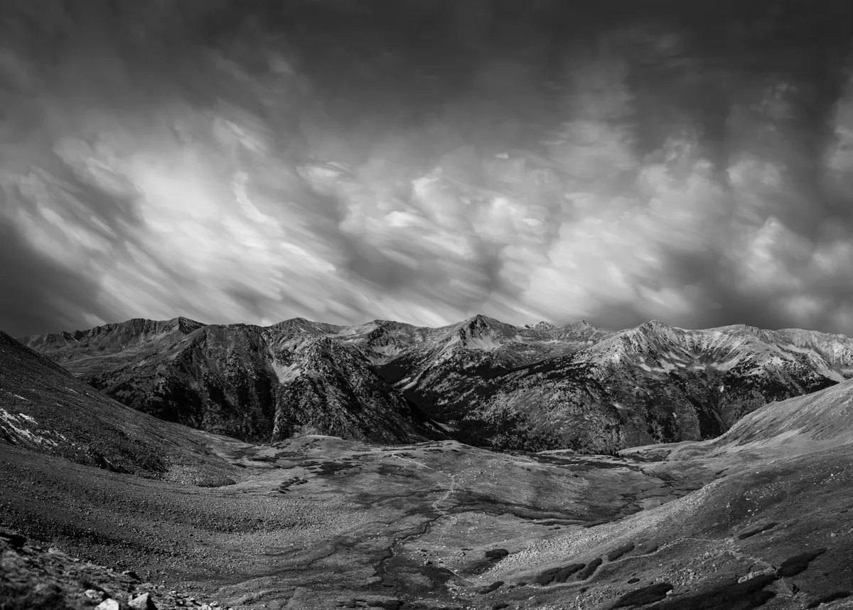 Black-and-white photograph of clouds streaked by a long exposure above a rugged mountain range and valley.