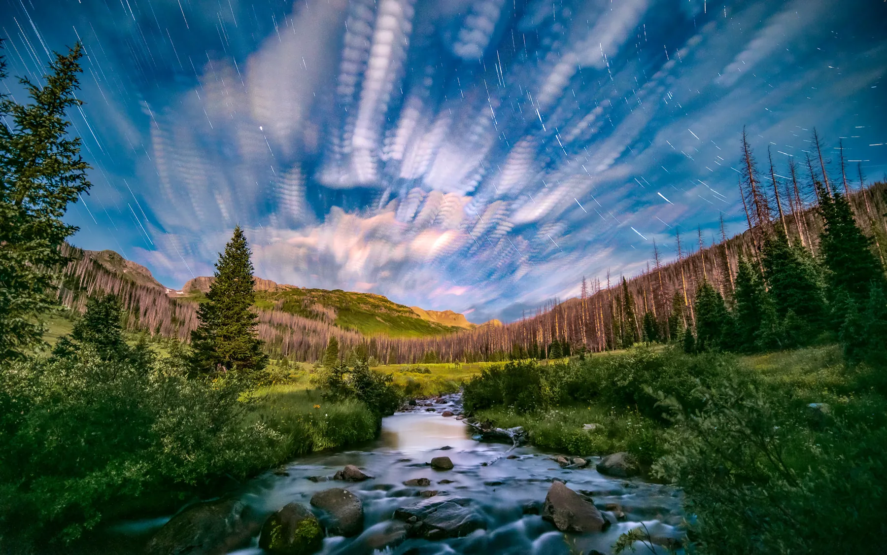 Night long exposure: clouds smeared into rippled streaks with faint star trails above a forested valley and a rocky creek.