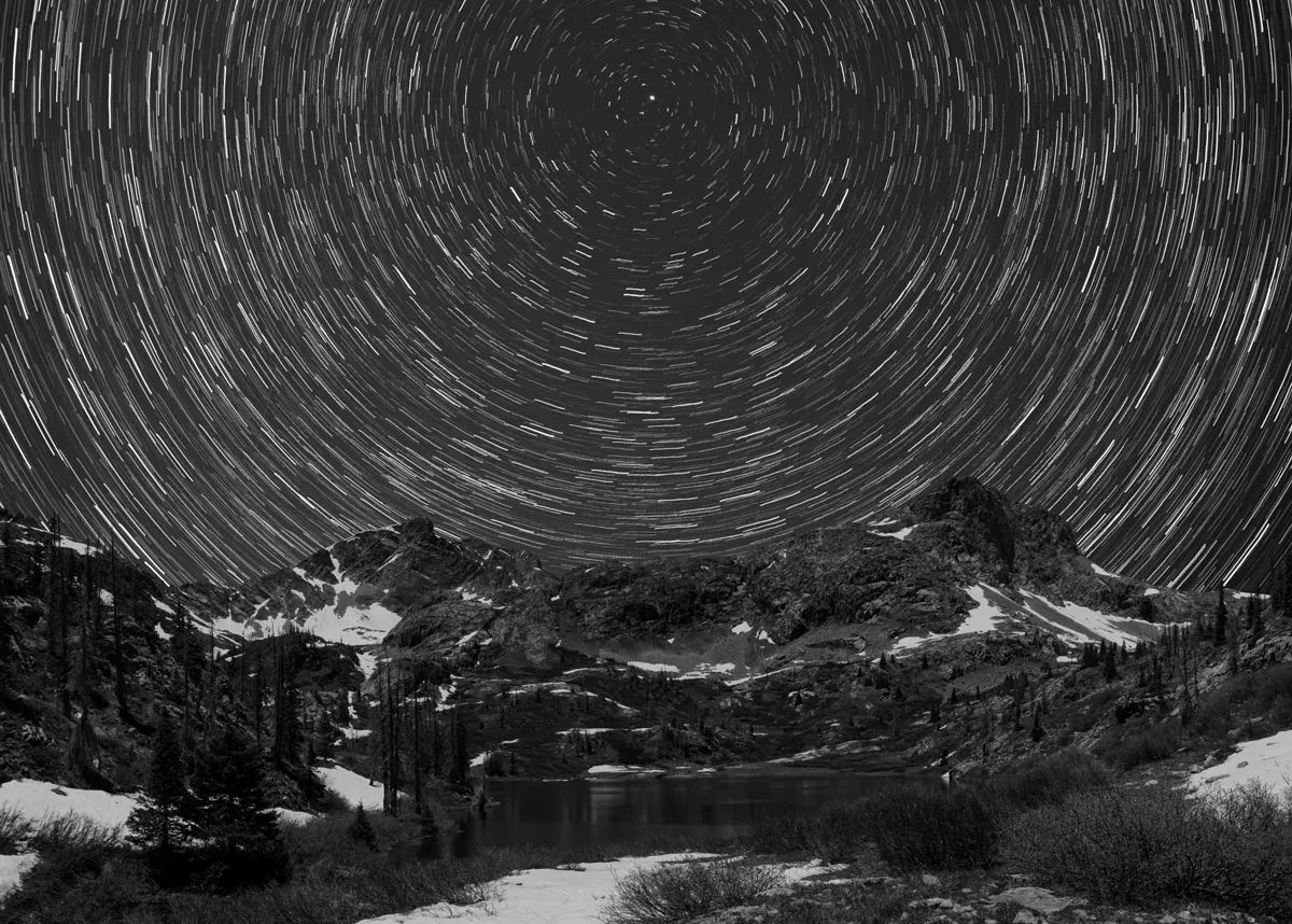 Black-and-white star trails in concentric circles over a snow-patched alpine lake and peaks.