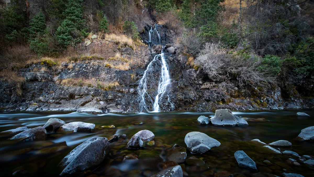 A small waterfall over dark rock into a river, the water blurred silky by a long exposure.